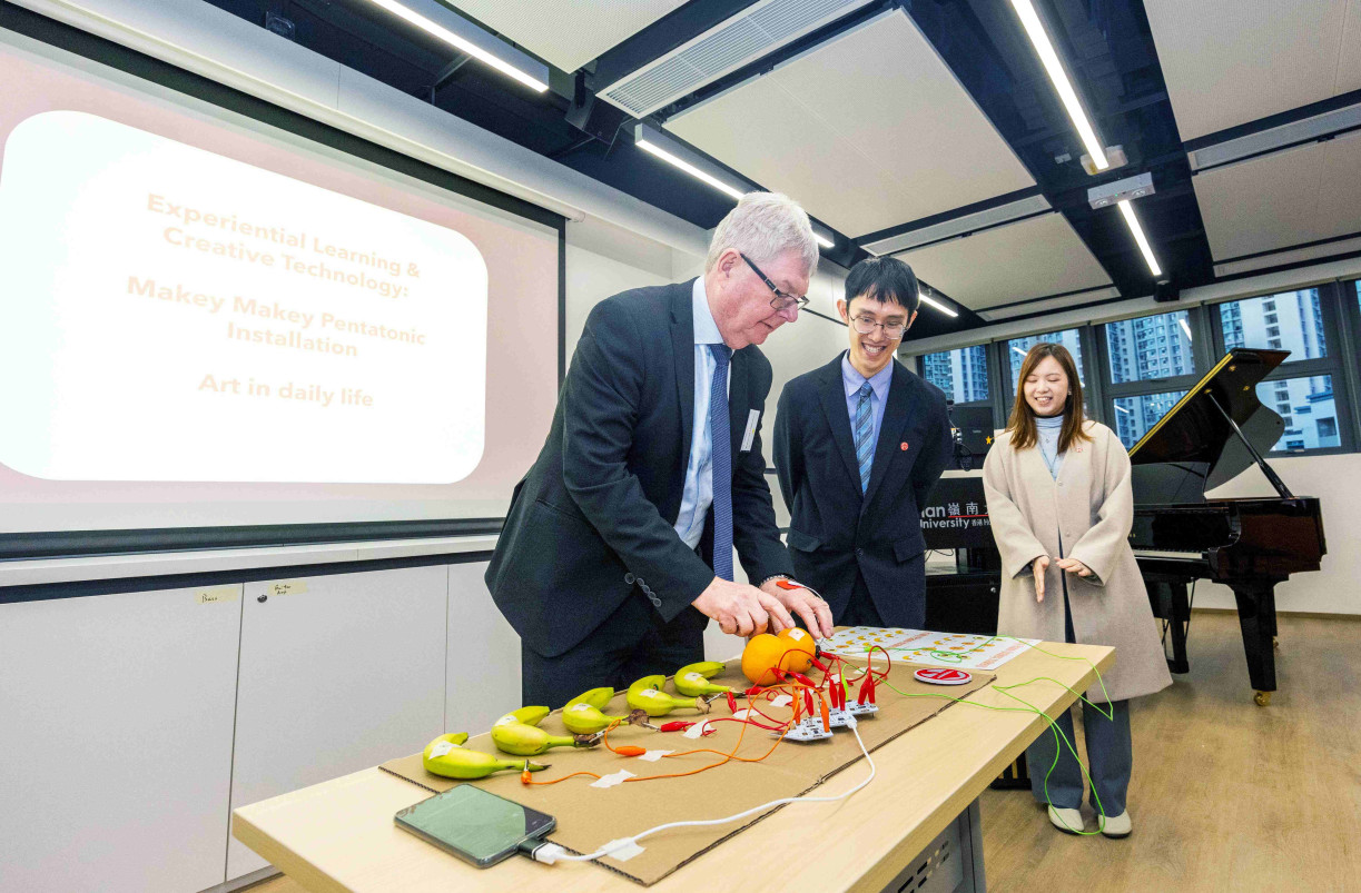 Prof David Cardwell (left), Member of the UGC, tries out the “Fruit Piano” using MaKey MaKey, connecting fruits to an electronic music device to create music through touch.