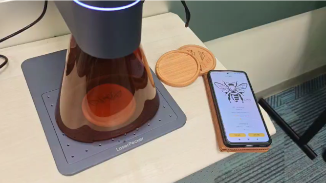 LaserPecker portable engraver working on a wooden coaster next to a smartphone design app.