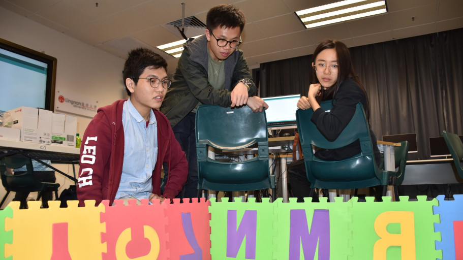  Students watching a project demonstration over a colorful foam puzzle tile barrier in a lab.