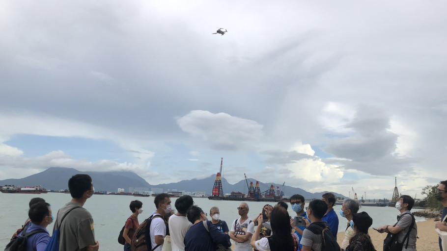  Workshop group watching a drone fly overhead on a coastal beach.